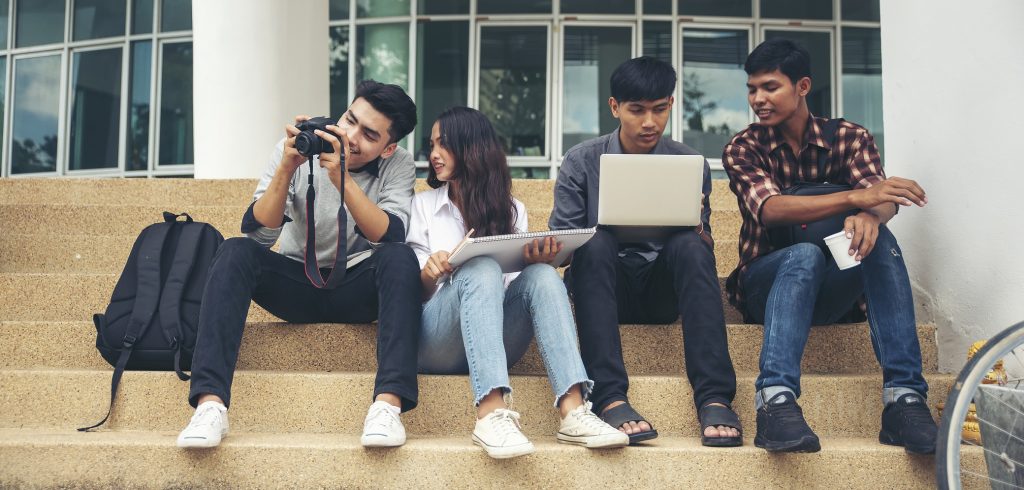 Four multi racial students sitting on the steps of a university library holding laptops, drawing pads, and a camera