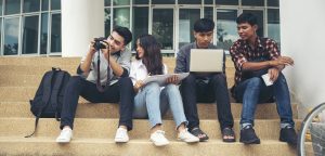 Four multi racial students sitting on the steps of a university library holding laptops, drawing pads, and a camera