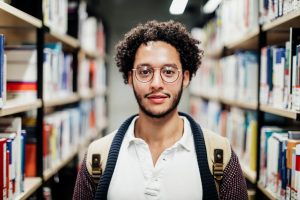 A portrait of a young man with a beard and glasses working in a public university library.