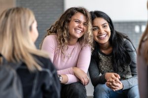 Two female young adults sit closely together, with their heads resting against each others, during a group therapy session. One is of African decent and the other is of Indian decent. Bother are dressed casually and are sitting among their multi-ethnic peers with smiles on their faces.