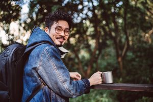 Young handsome asian man smiling joyfully at camera while drinking with his reusable coffee cup in country park