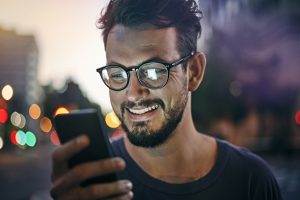 Young man reading text message while wearing glasses and standing outside at dusk